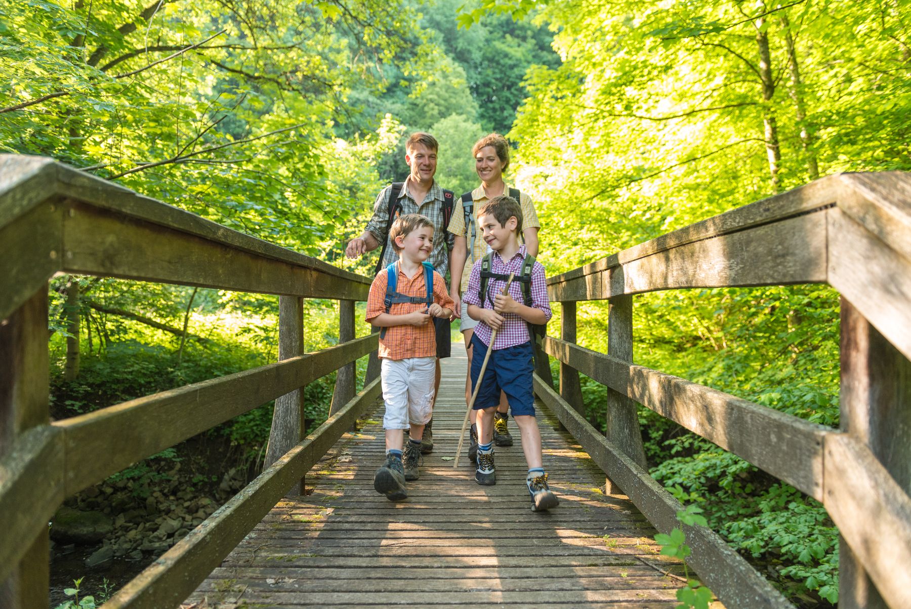A family of four, two adults and two children, walk together on a wooden bridge in a lush, green forest. The children are smiling and carrying walking sticks, and everyone appears to be enjoying a hike.